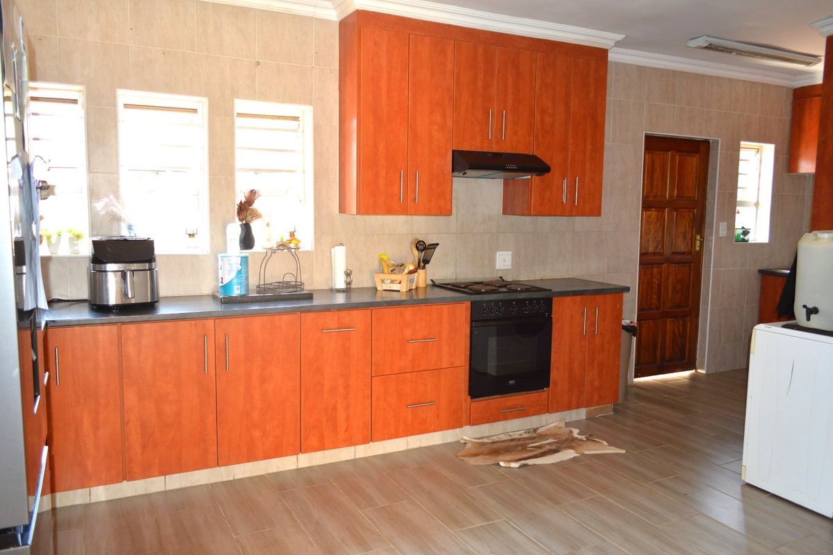 A functional kitchen featuring warm-toned wooden cabinetry, built-in oven and hob, tiled splashbacks, and durable floor tiles with direct access to an adjacent room.