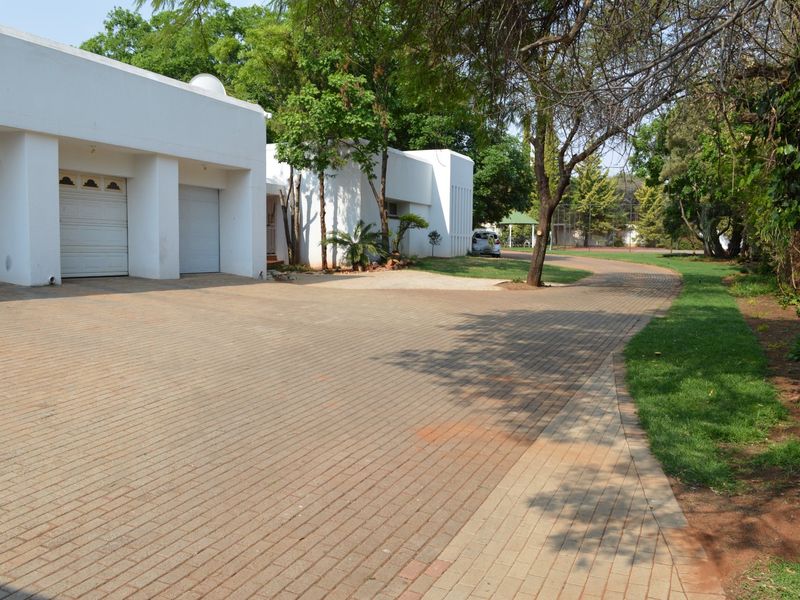 Spacious front exterior featuring a large paved driveway leading to a triple garage, surrounded by a well-established garden with mature trees.