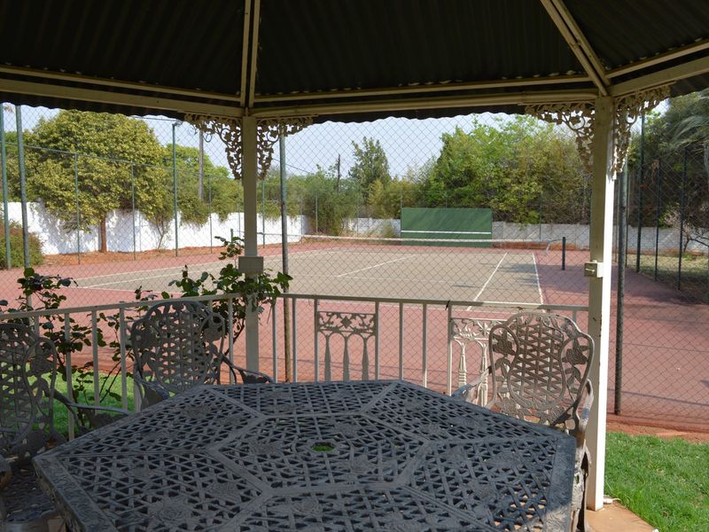 A private tennis court with a practice wall, viewed from a shaded gazebo seating area.