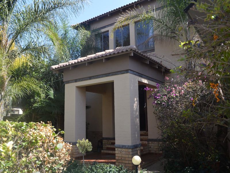 A well-maintained double-storey house entrance featuring a covered porch with brick-based pillars and terracotta tiling, surrounded by established garden landscaping.