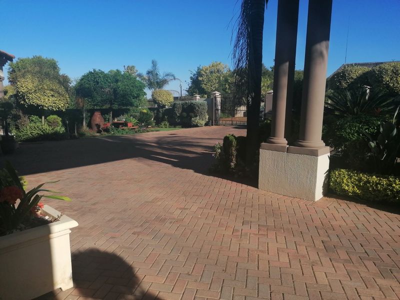 A well-maintained front entrance area featuring a brick paved driveway, decorative structural columns, and established garden landscaping leading to a secure gated entrance.