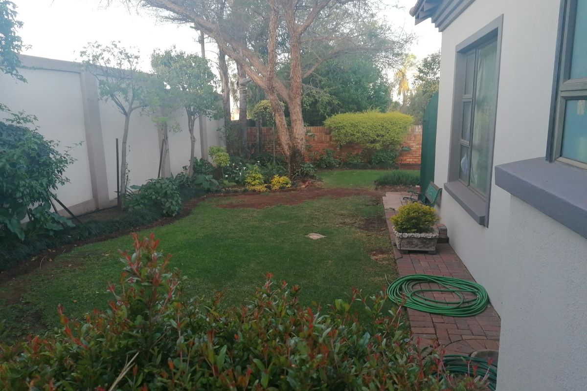 A well-maintained rear garden featuring a manicured lawn, established trees, and brick-paved pathways alongside the property's exterior wall.