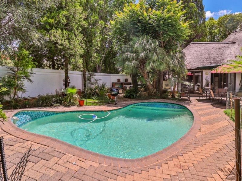 A private backyard featuring a kidney-shaped swimming pool, brick-paved patio area, and a built-in braai, set against a lush garden and thatched-roof structure.