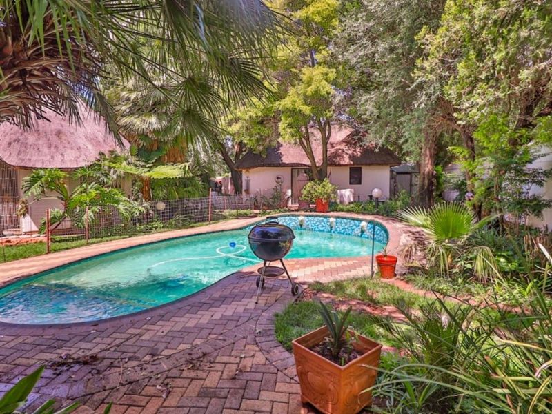 A private outdoor pool area featuring a curved swimming pool surrounded by brick paving, mature garden landscaping, and traditional thatched roof outbuildings.