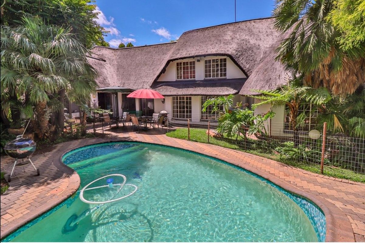 Rear view of the property featuring a thatched roof, a swimming pool, and a paved patio area surrounded by established garden landscaping.