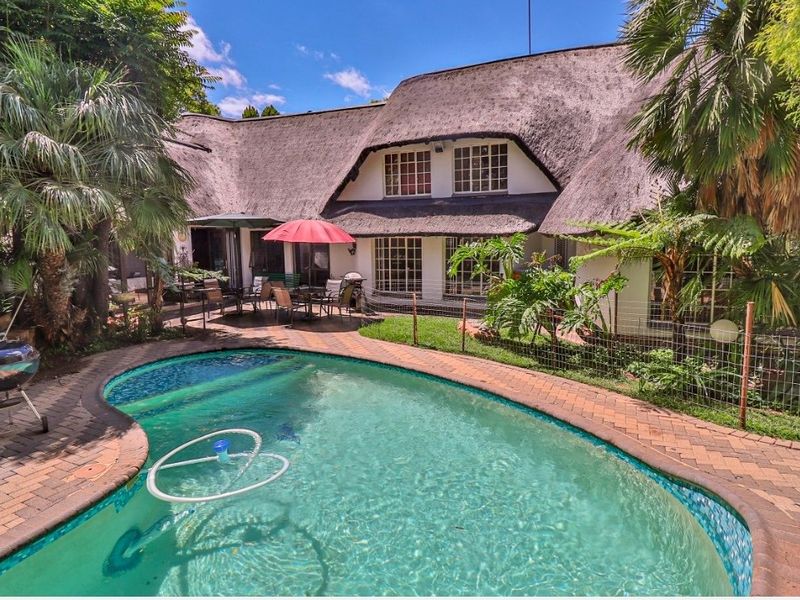 Rear view of the property featuring a thatched roof, a swimming pool, and a paved patio area surrounded by established garden landscaping.
