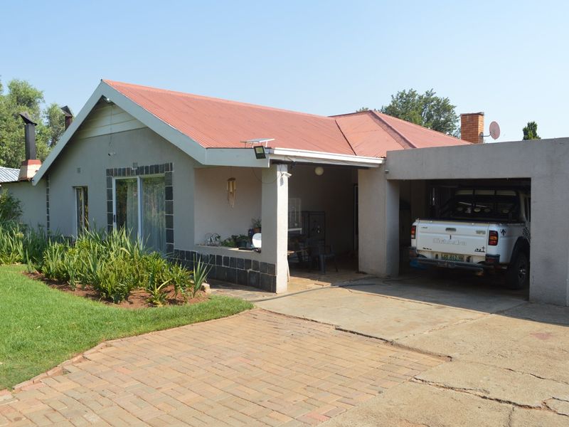 Front view of a single-story house featuring a corrugated metal roof, stone cladding accents, a covered carport, and a paved driveway.