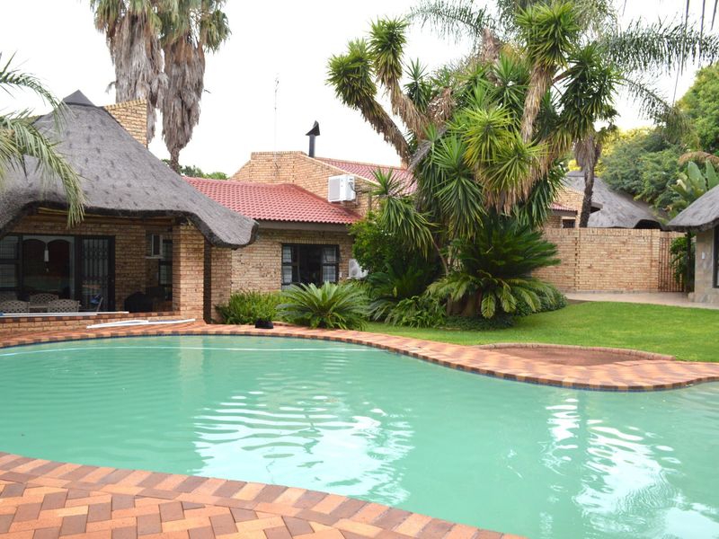 A well-maintained outdoor pool area featuring a curved swimming pool, brick paving, and a thatched roof structure set within a landscaped garden.