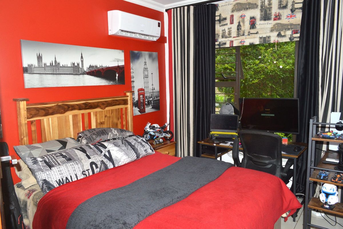 A bedroom featuring a bold red accent wall, a split-system air conditioning unit, and a large window overlooking garden greenery.