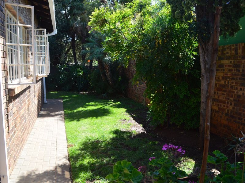 A private rear garden area featuring a brick-paved walkway alongside the townhouse exterior wall, mature trees, and a brick boundary wall.
