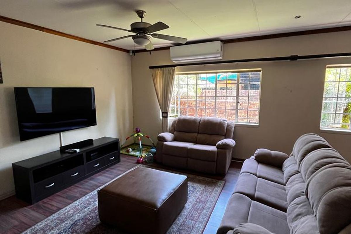 A comfortable living room featuring laminate flooring, a ceiling fan, and a wall-mounted air conditioning unit, with natural light from grid-pattern windows.