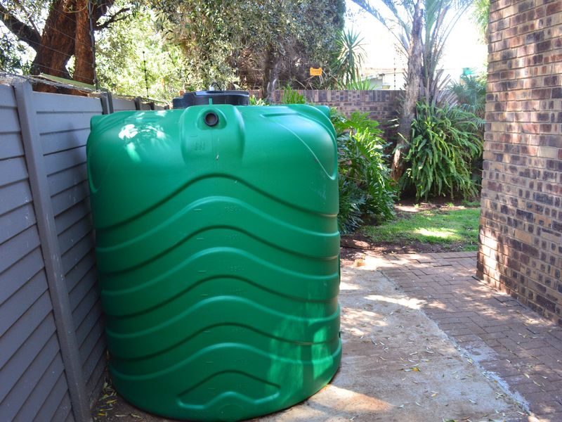 A green water storage tank installed in the side garden area, featuring brick boundary walls and paved access.