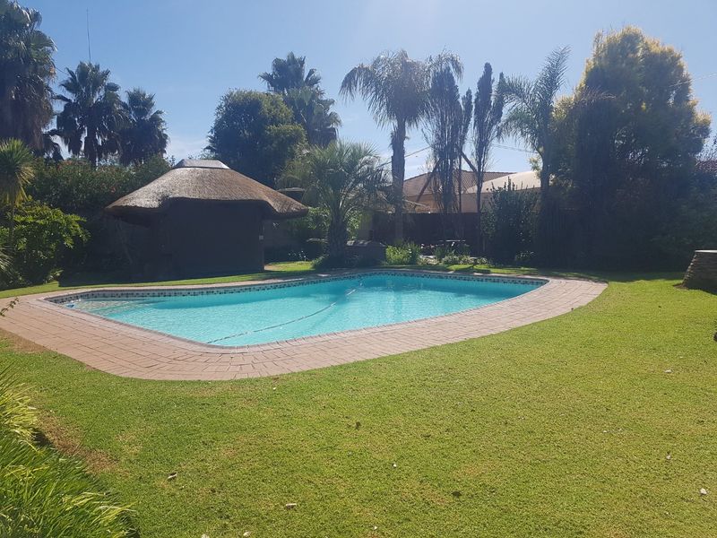 A well-maintained swimming pool area featuring a paved surround, a traditional thatched lapa, and established garden landscaping.