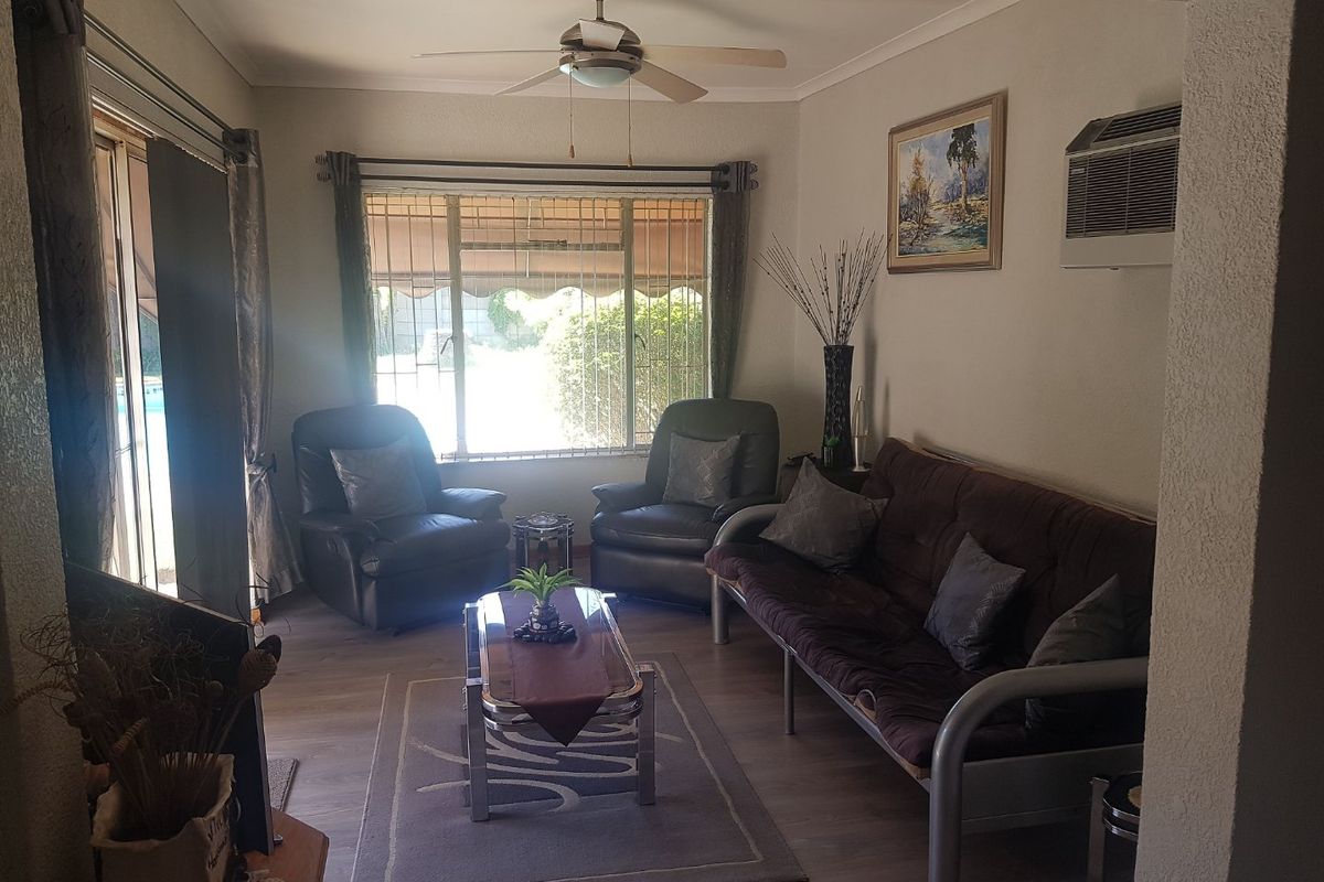 A comfortable living room featuring laminate flooring, a ceiling fan, wall-mounted air conditioning, and large windows with security bars overlooking the exterior.