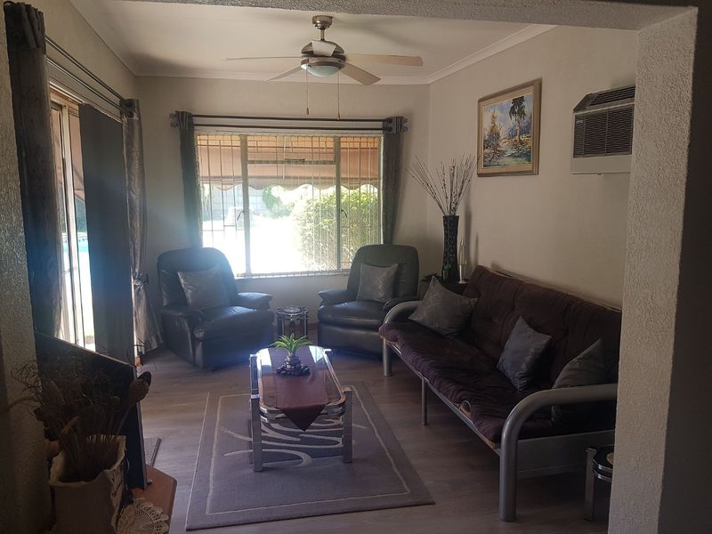 A comfortable living room featuring laminate flooring, a ceiling fan, wall-mounted air conditioning, and large windows with security bars overlooking the exterior.