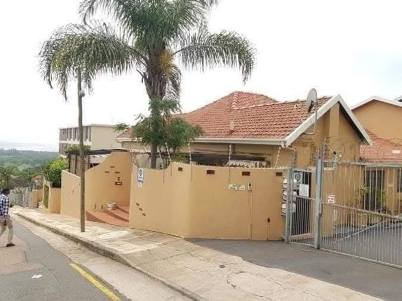 Front exterior view of a townhouse featuring a tiled roof, perimeter wall with security fencing, and a gated driveway entrance.