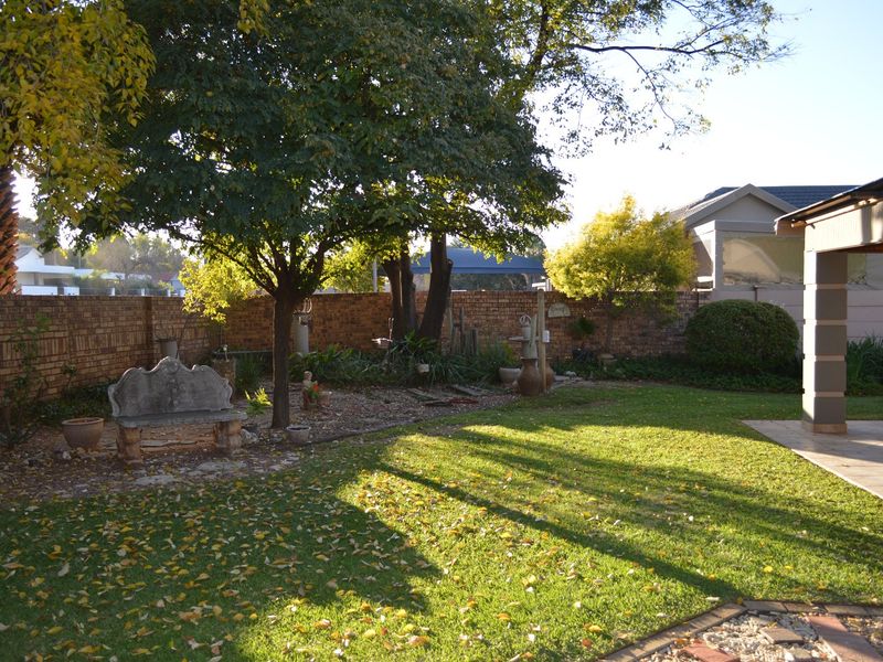 A well-maintained private garden featuring a lush lawn, mature trees, and a brick boundary wall, adjacent to a paved patio area.