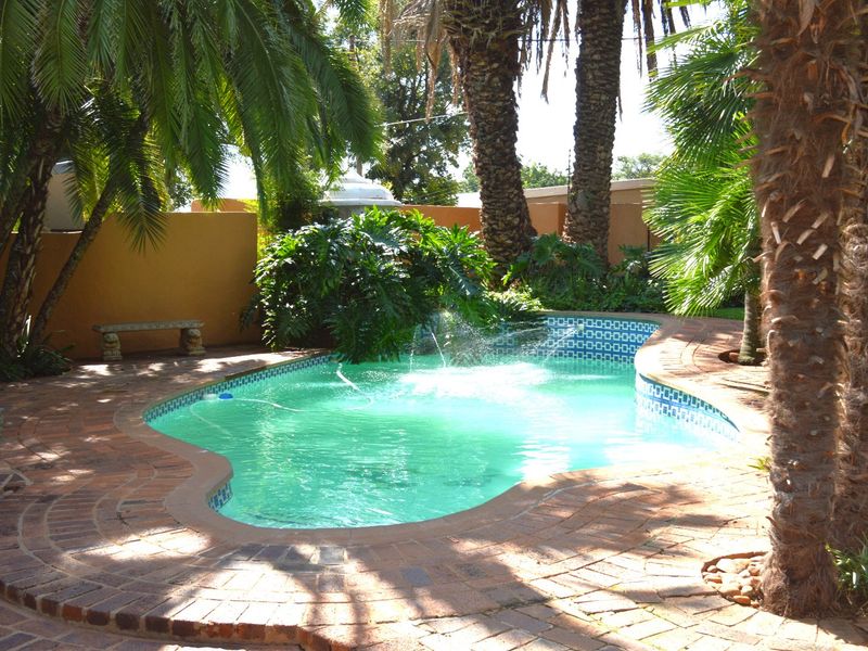 A well-maintained swimming pool area featuring brick paving, mature palm trees, and a surrounding boundary wall in a lush garden setting.
