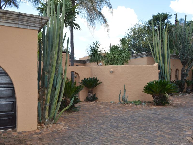 A well-maintained front exterior featuring a paved driveway, stucco walls, and an arched wooden entrance door surrounded by mature desert-style landscaping.