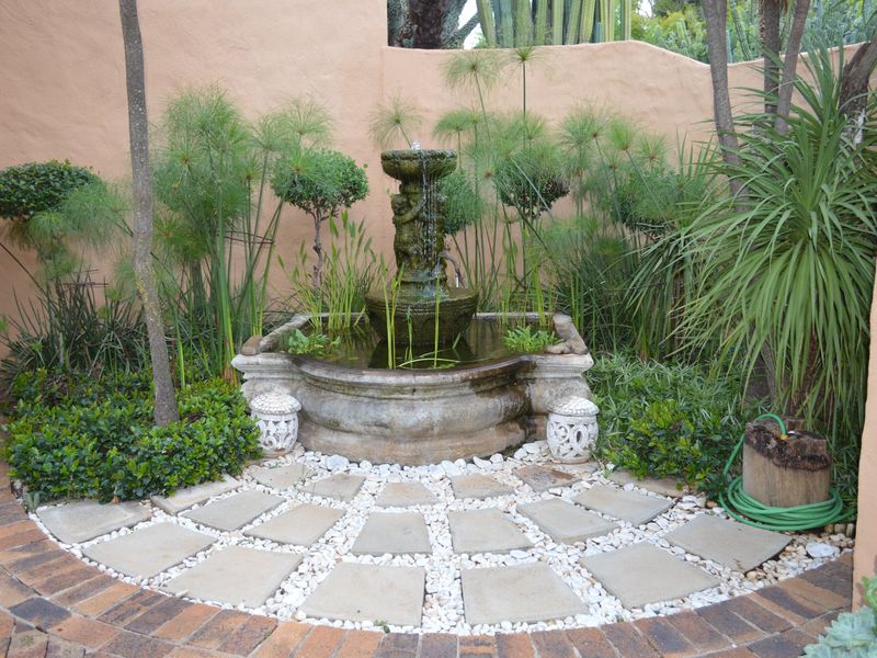 A tranquil garden corner featuring a central stone fountain, surrounded by lush greenery and a paved circular patio area.