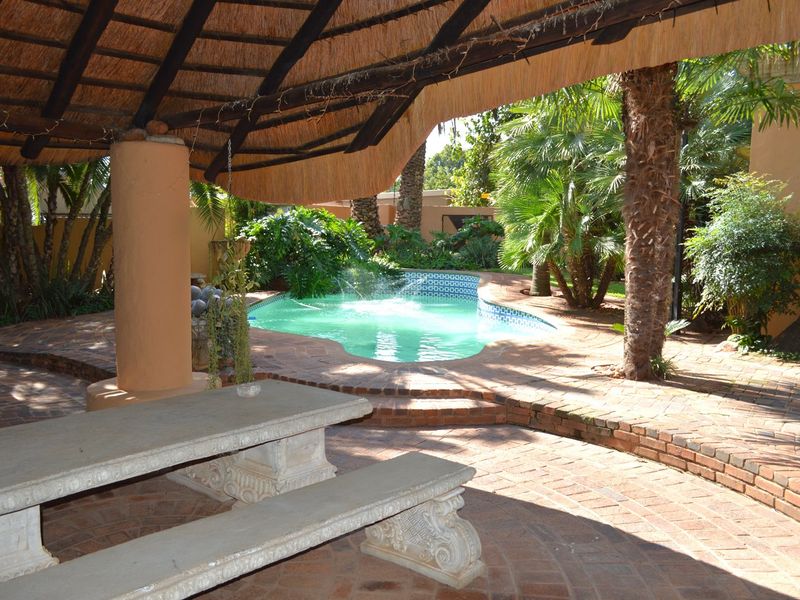 Outdoor entertainment area featuring a thatched lapa, brick-paved patio, and a swimming pool with a built-in fountain, set within a landscaped garden.