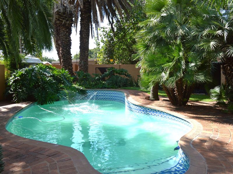 A landscaped backyard featuring a curved swimming pool with a built-in water feature, surrounded by mature palm trees and brick paving.