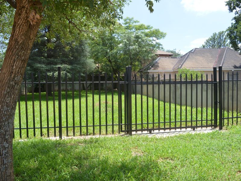A view of a vacant residential plot secured with a black metal picket fence and gate, featuring a well-maintained lawn.