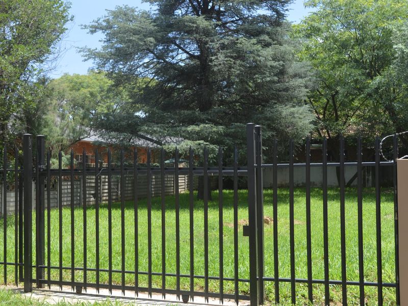 A vacant residential plot secured with black metal fencing and a gate, featuring a well-maintained lawn and mature trees.