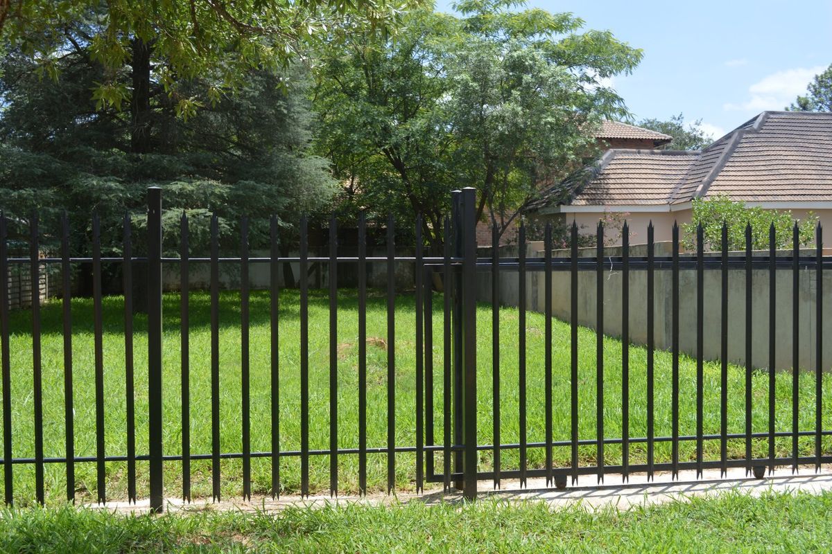 A view of a vacant residential plot featuring a secure black metal spike fence, pedestrian gate, and established lawn area.