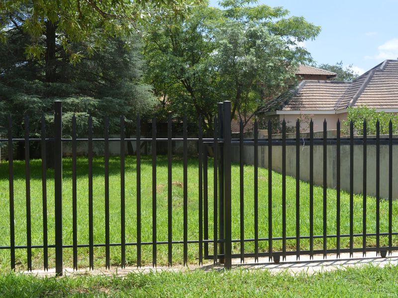 A view of a vacant residential plot featuring a secure black metal spike fence, pedestrian gate, and established lawn area.