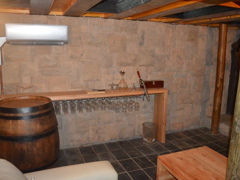 A rustic-style bar area featuring stone-clad walls, exposed timber ceiling beams, slate flooring, and a custom-built timber counter with integrated glass storage.