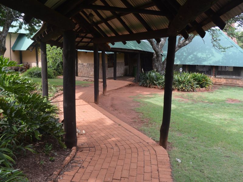 A covered brick-paved walkway leads through a landscaped garden area, connecting sections of the farm-style dwelling with timber posts and corrugated roofing.