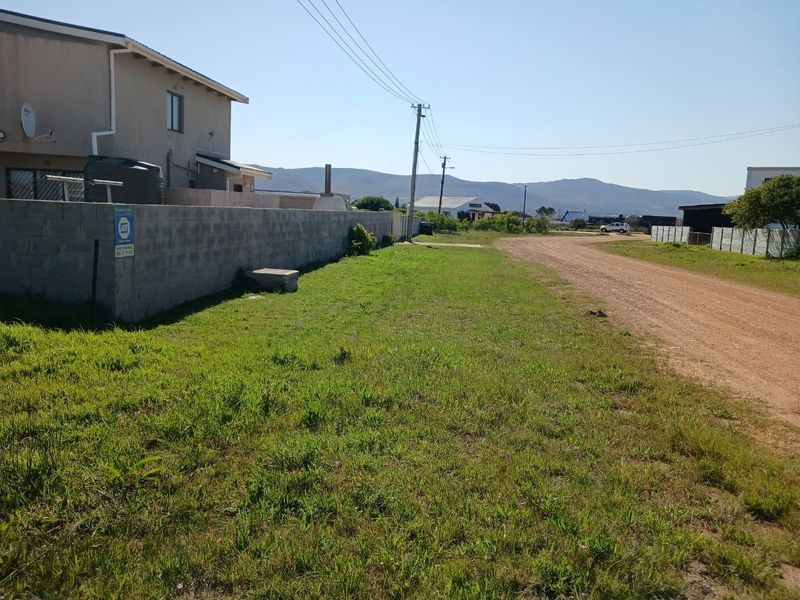 A street-level view of a vacant residential plot featuring an unpaved road, boundary wall, and surrounding neighborhood infrastructure.