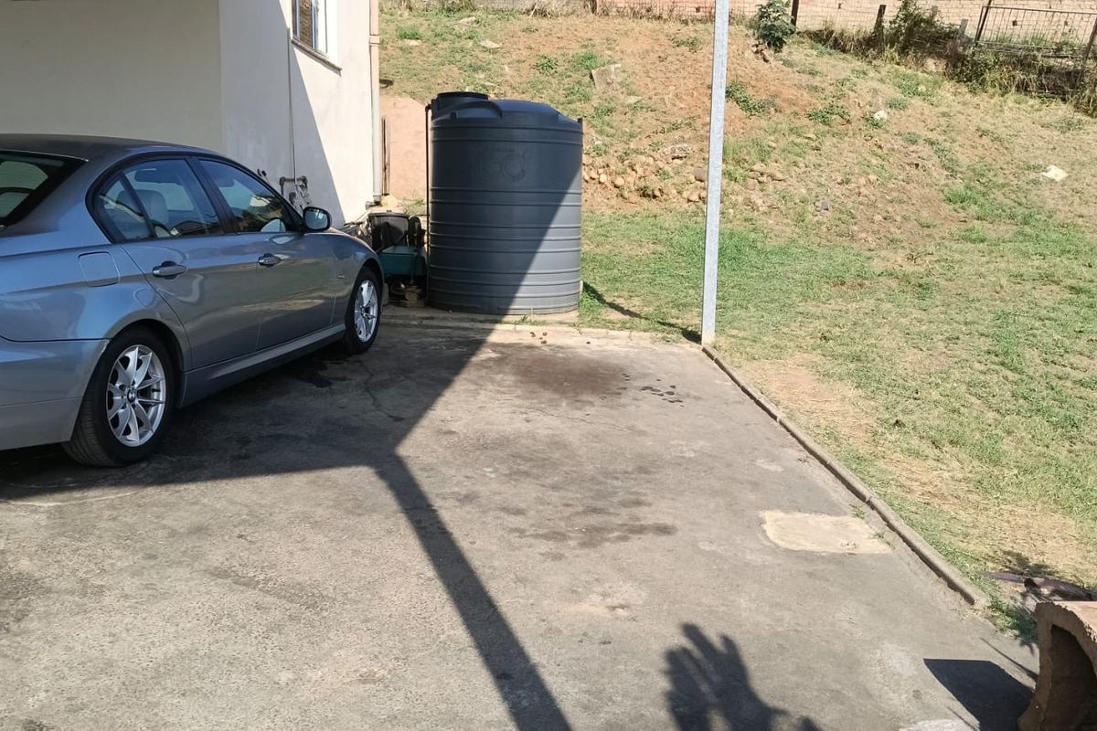 Rear exterior area featuring a concrete driveway, covered parking space, and a large water storage tank set against a sloped grass yard.