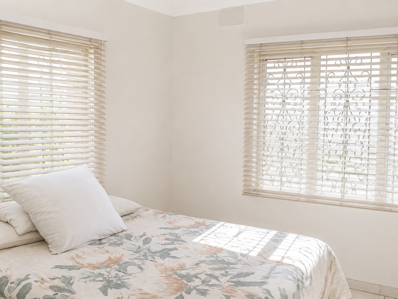 A bright bedroom featuring neutral walls, tiled flooring, and windows fitted with venetian blinds and security bars.