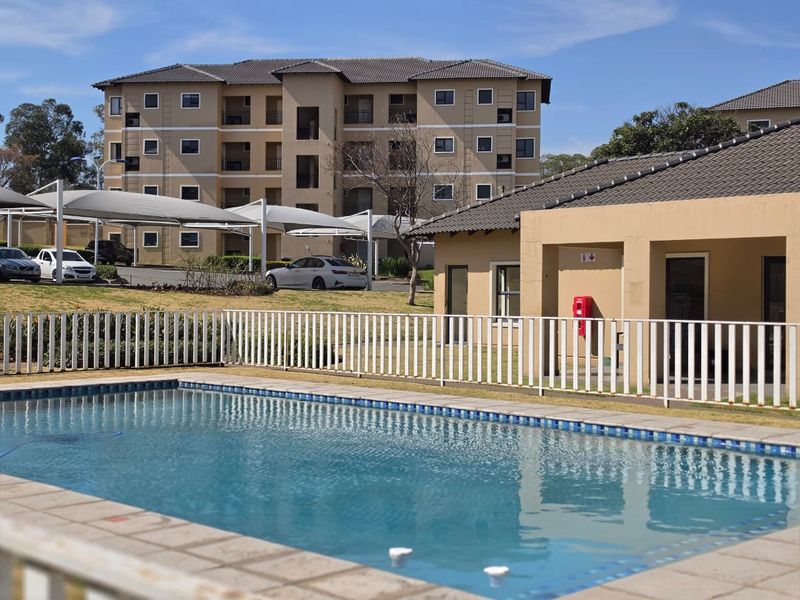 A well-maintained communal swimming pool area featuring tiled coping and secure metal fencing, set within a modern apartment complex with landscaped grounds.