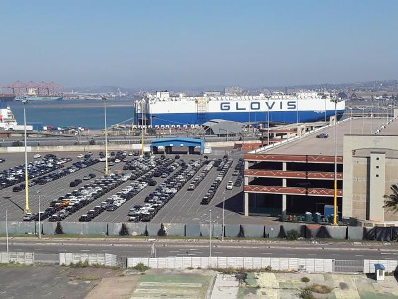 An elevated view overlooking a busy industrial harbour, featuring a large cargo ship and extensive vehicle storage facilities.