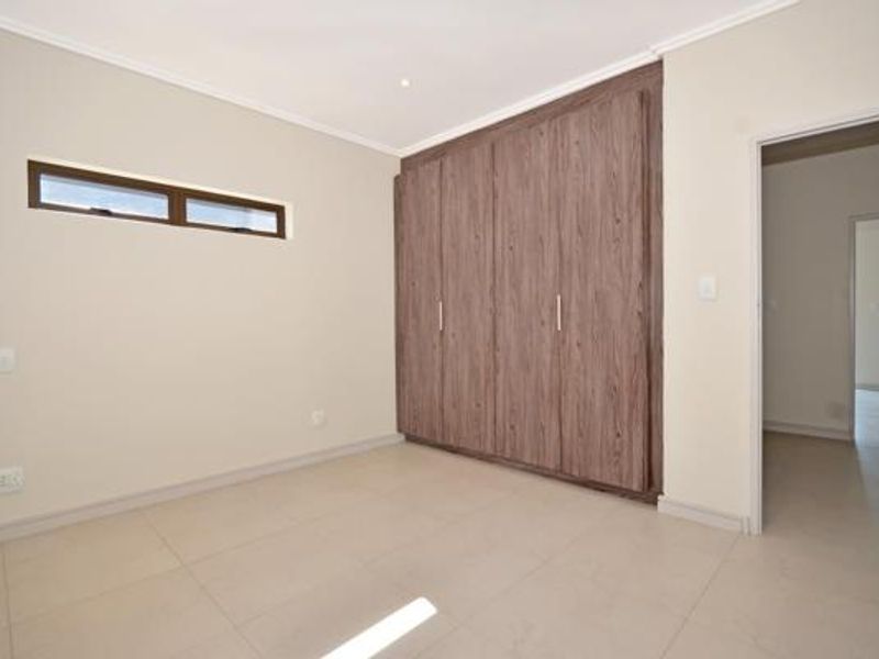 A bright, empty bedroom featuring neutral tiled flooring, built-in wooden cupboards, and a high-set clerestory window.