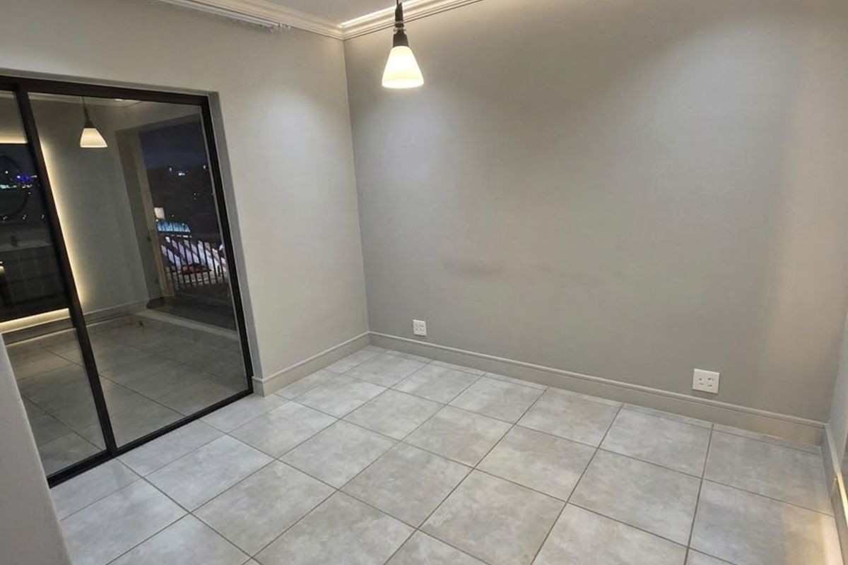 A bright, modern, and empty living area featuring neutral tiled floors, recessed ceiling lighting, cove lighting, and a large sliding glass door leading to a balcony.