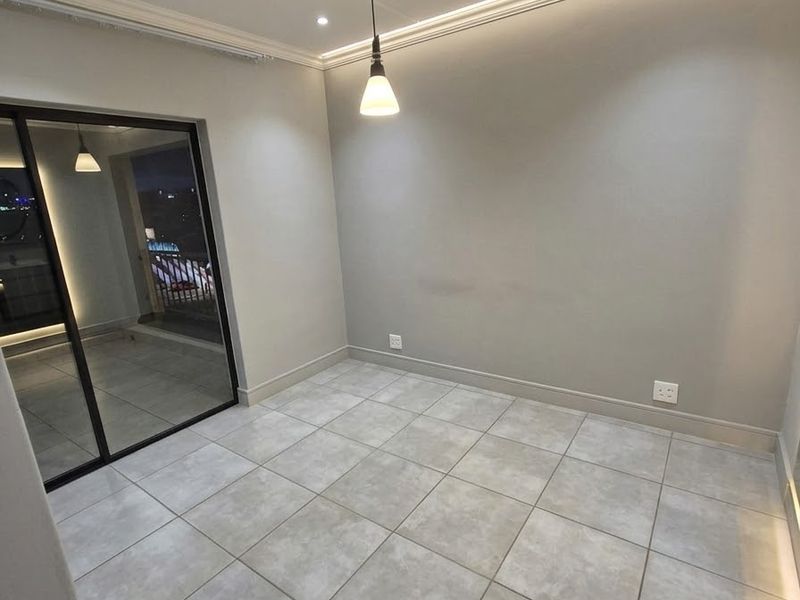 A bright, modern, and empty living area featuring neutral tiled floors, recessed ceiling lighting, cove lighting, and a large sliding glass door leading to a balcony.