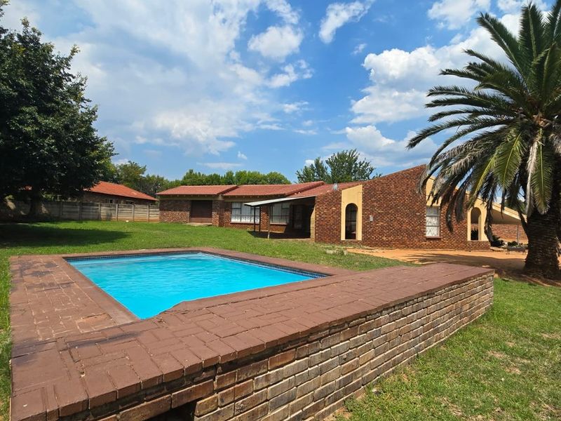 A spacious backyard featuring a raised brick swimming pool, established lawn, and a face brick house exterior under a clear blue sky.