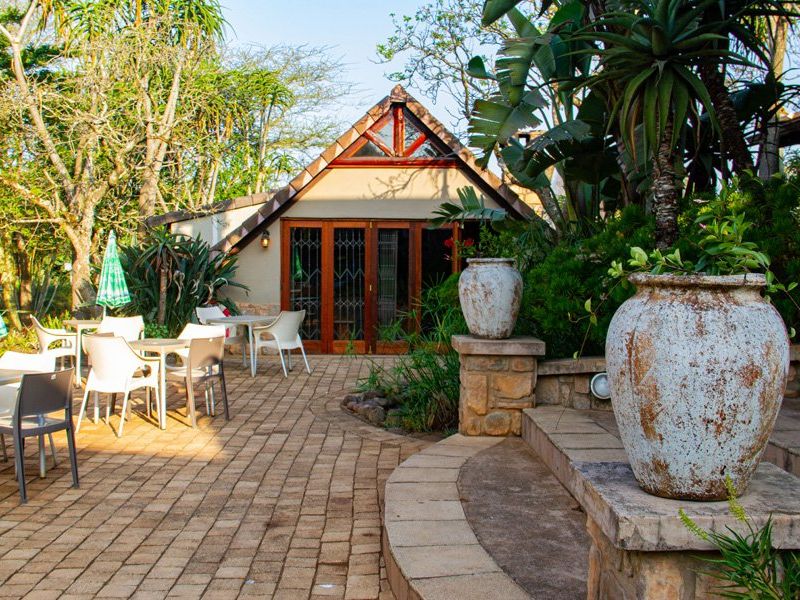 A well-maintained paved outdoor patio area featuring stone retaining walls, lush garden surroundings, and access to the building via timber-framed glass doors.