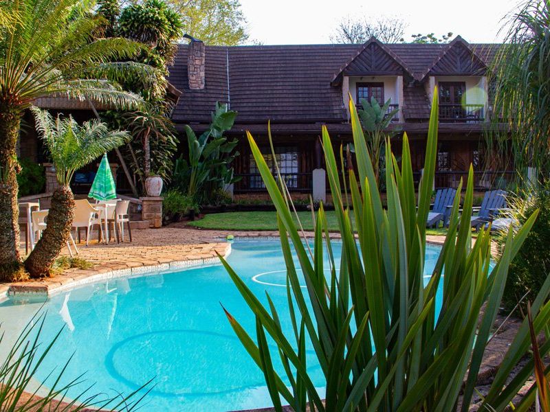 A well-maintained outdoor pool area featuring a paved patio, lush landscaping, and a view of the multi-story building with balconies and a tiled roof.