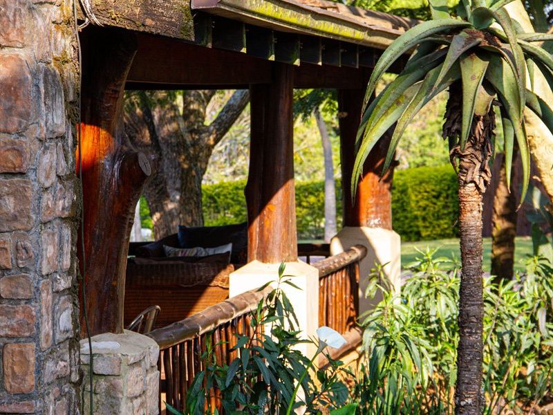 A rustic covered patio area featuring natural stone cladding, timber log support columns, and a timber railing overlooking a garden setting.