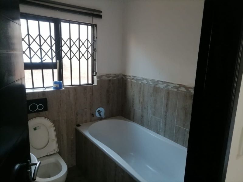 A functional bathroom featuring a built-in bathtub, wall-mounted toilet, and modern tiled wall finishes with a security-fitted window.