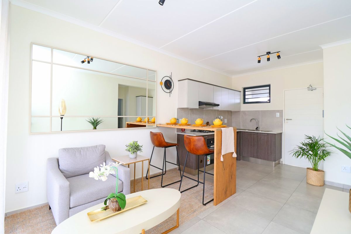 Modern open-plan living area featuring a compact kitchen with built-in cabinetry, integrated appliances, and a breakfast bar, finished with neutral tiled flooring.