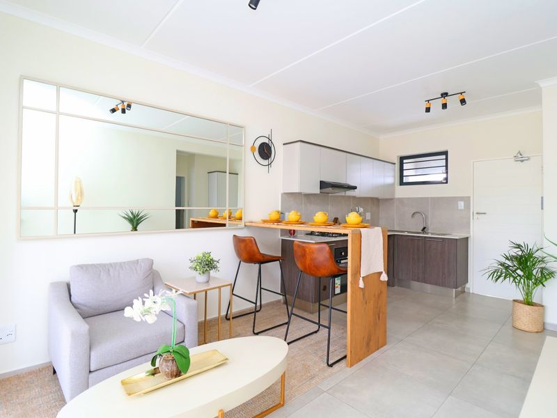 Modern open-plan living area featuring a compact kitchen with built-in cabinetry, integrated appliances, and a breakfast bar, finished with neutral tiled flooring.