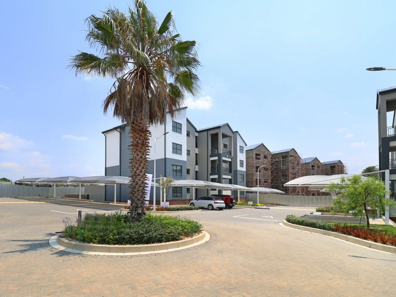 Exterior view of a modern apartment complex featuring paved driveways, covered parking bays, and landscaped garden areas with ongoing development in the background.