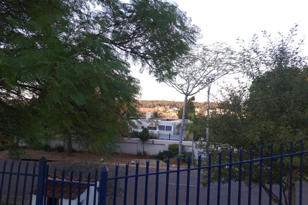 A street-level view of the property frontage, featuring established trees, a boundary wall, and metal perimeter fencing.
