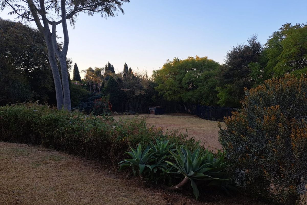 A spacious residential garden featuring mature trees, established hedging, and a well-defined lawn area enclosed by perimeter fencing.